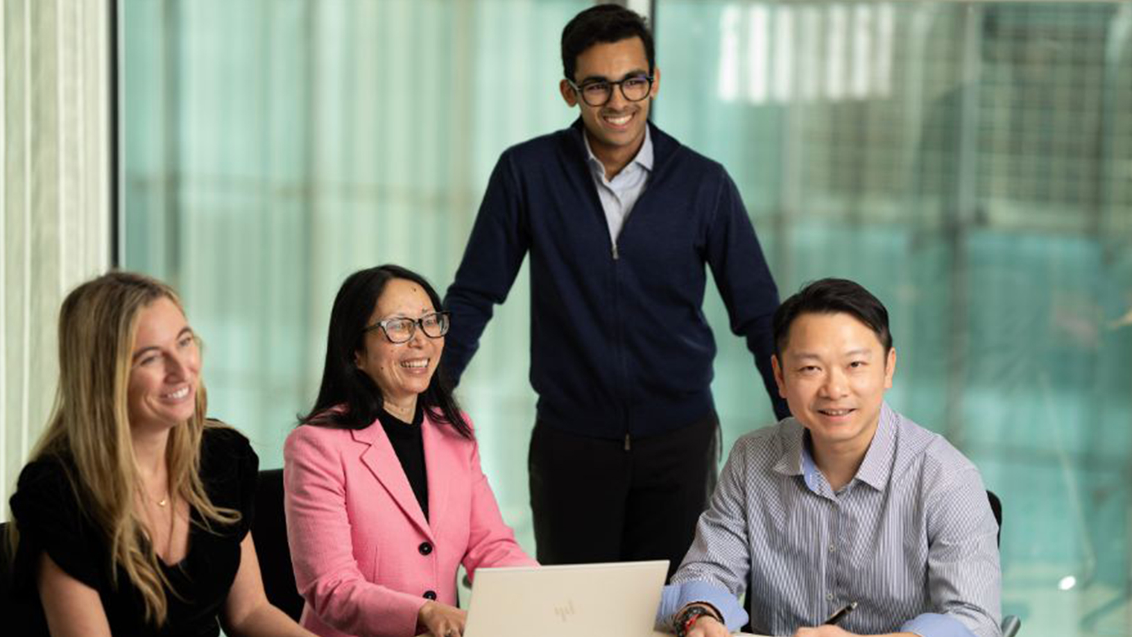 Four coworkers smiling in a glass-walled office, gathered around a laptop during a meeting. on Life At Fitch