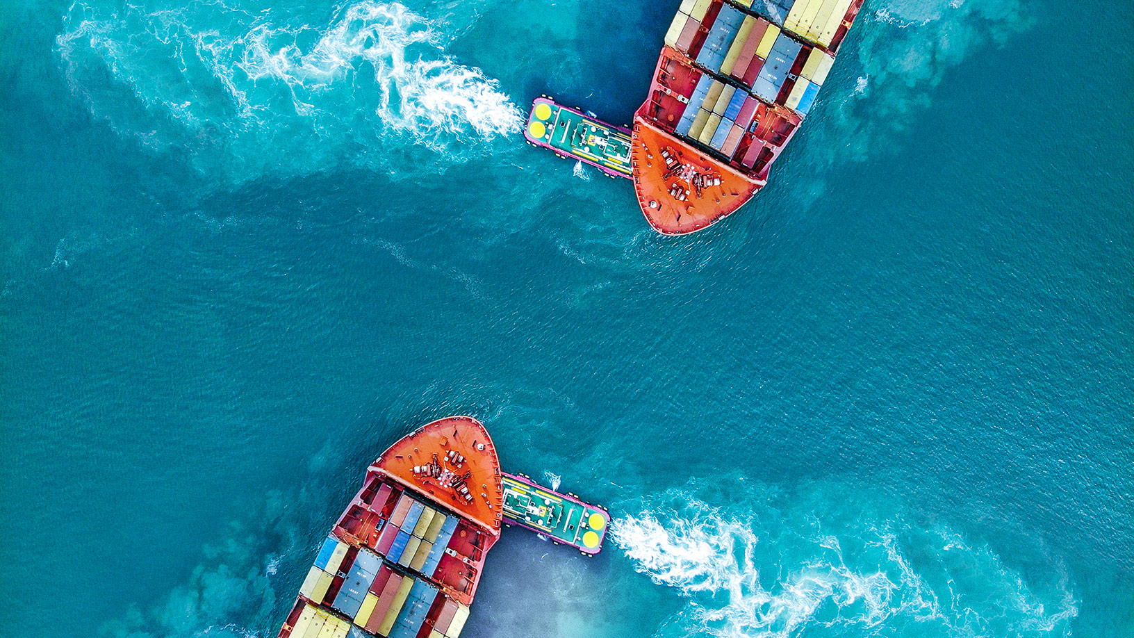 Aerial view of two container ships in turquoise water, each guided by a tugboat leaving white wakes. on Fitch Ratings: Global Economic Outlook