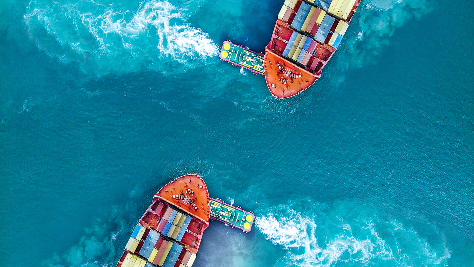 Aerial view of two container ships in turquoise water, each guided by a tugboat leaving white wakes. on Fitch Ratings: Global Economic Outlook