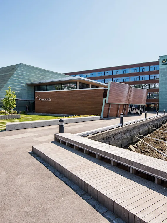 A modern building with glass and wood facades next to a waterfront promenade and a wooden pier, with mountains in the background. Text: Sjohuset.