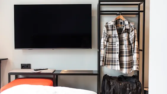 A plaid shirt hangs on a rack beside a black duffel bag; a flat-screen TV is on the wall in a minimalistic room.