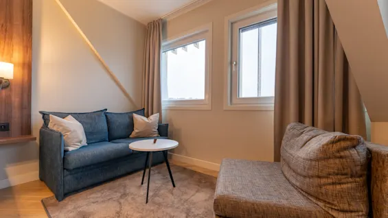 Et koselig oppholdsrom med en blå sofa, et lite rundt bord og en annen komfortabel sitteplass, badet i naturlig lys fra to vinduer.