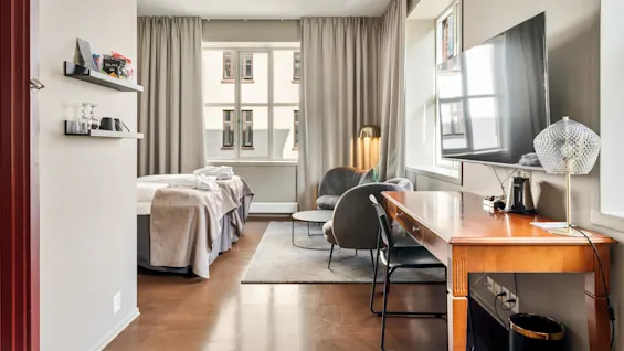 Et lyst hotelværelse med to enkeltsenge med gråt sengetøj, et vægmonteret fjernsyn, et træskrivebord med en lampe og lænestole nær et vindue skaber en hyggelig atmosfære.