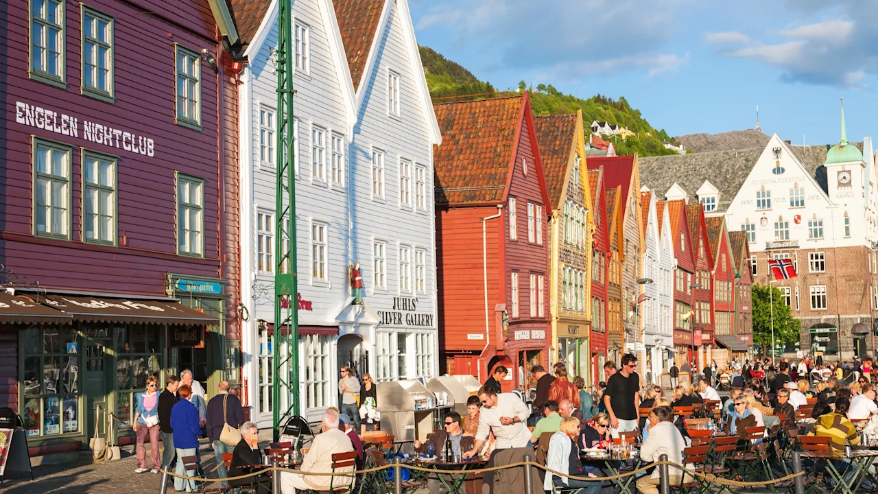 Colorful wooden buildings line a bustling street as people sit at outdoor cafes. Signs read "ENGELEN NIGHTCLUB" and "JULES DRIVER GALLERY."