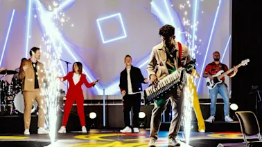 Live band på scenen med sangere, gitarist og en person foran bildet som spiller keytar, med gnister på scenen
