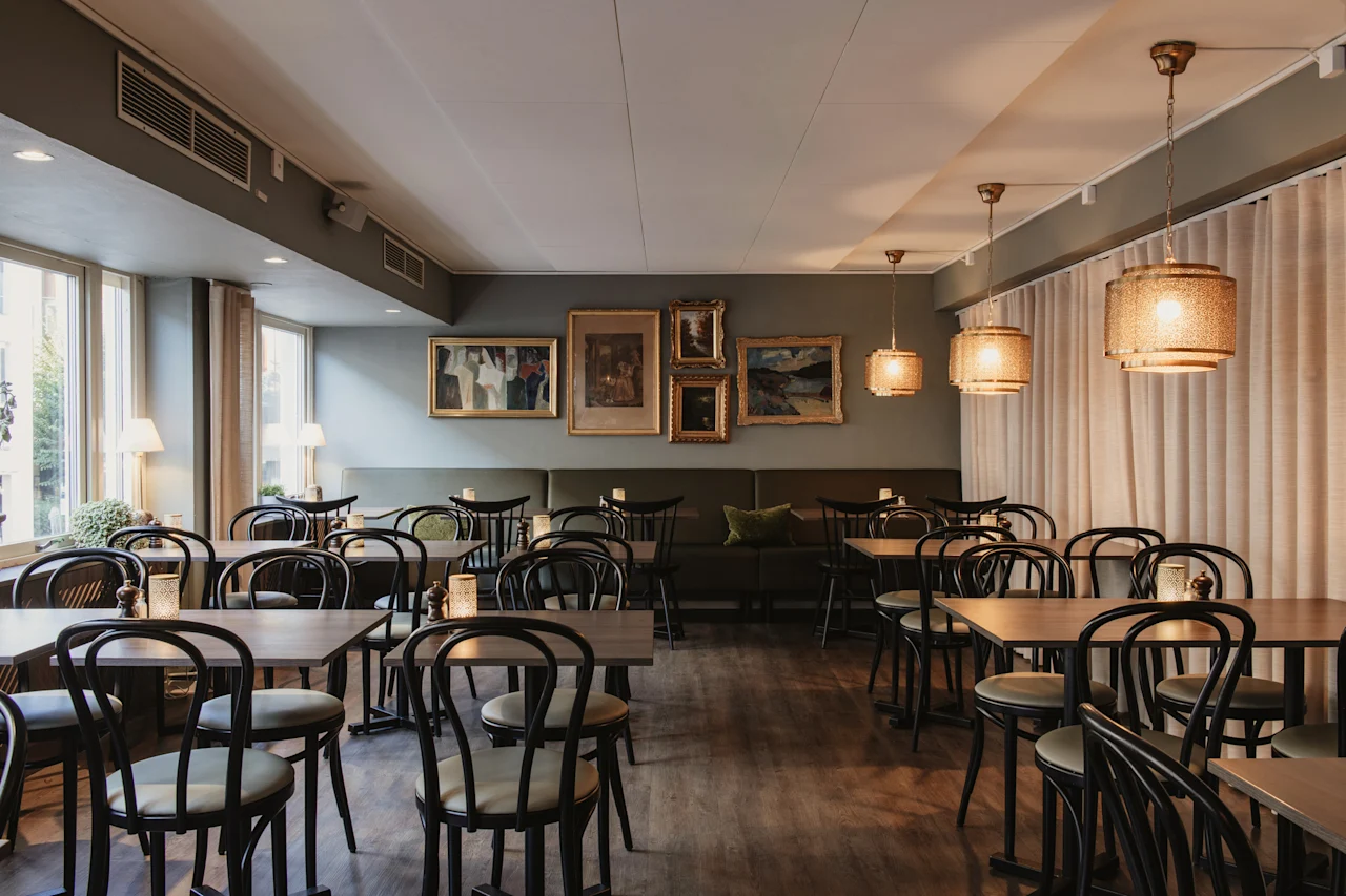 En mysig restaurangmatsal med flera bord och stolar, upplyst av hängande lampor och naturligt ljus från fönster.