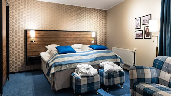 Ett välbäddat säng med blått och vitt lakan i ett välbelyst hotellrum, omgiven av två rutiga fåtöljer och väggkonst.