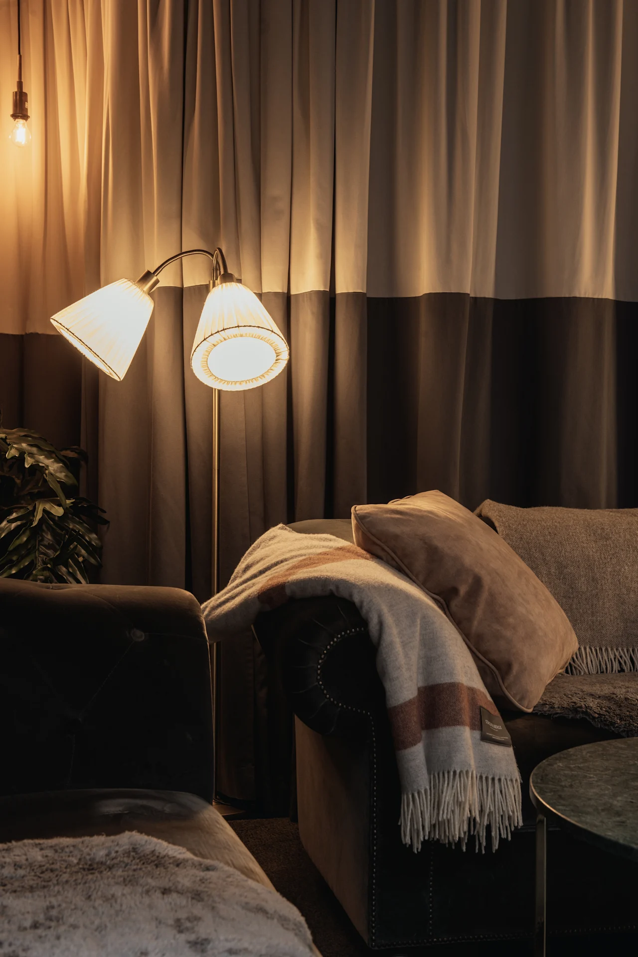 Et hyggeligt opholdsrum med varm belysning fra en gulvlampe, en komfortabel sofa med puder og et tæppe samt bløde gardiner.