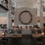 Matbord är ordnade med tända ljus i en modern, stilren restaurang. En stor dekorativ krans pryder en central vit vägg, omgiven av moderna sittplatser och belysningsarmaturer.
