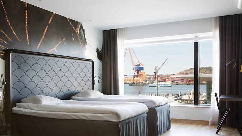 Two neatly made twin beds positioned beside a large window overlook a harbor scene, where cranes and a ship are visible. The room has a modern decor with wall art.