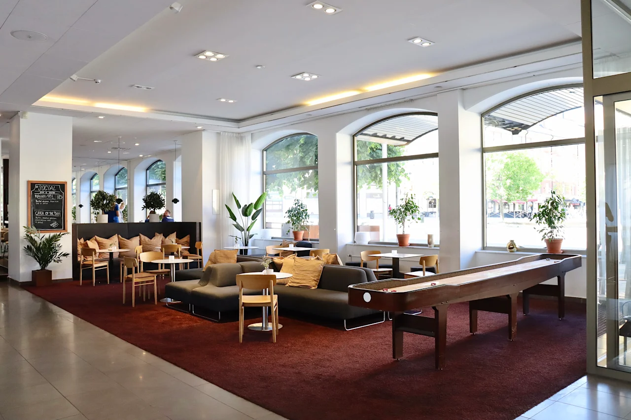 Lounge area featuring sofas and chairs on a red carpet, with plants and large windows overlooking the street. A shuffleboard table completes the modern, cozy interior.