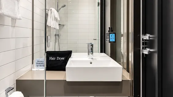 Modern hotel bathroom featuring a white sink, large mirror, tiled walls, and a bag labeled Hair Dryer.
