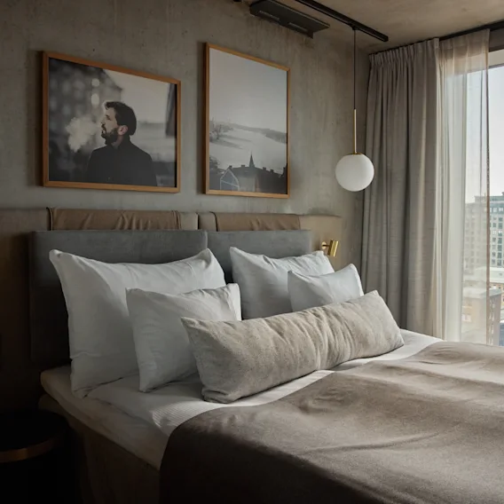 Ett mysigt hotellrum med en bäddad säng, mjuka kuddar och konst på väggen. Ett stort fönster erbjuder utsikt över staden.