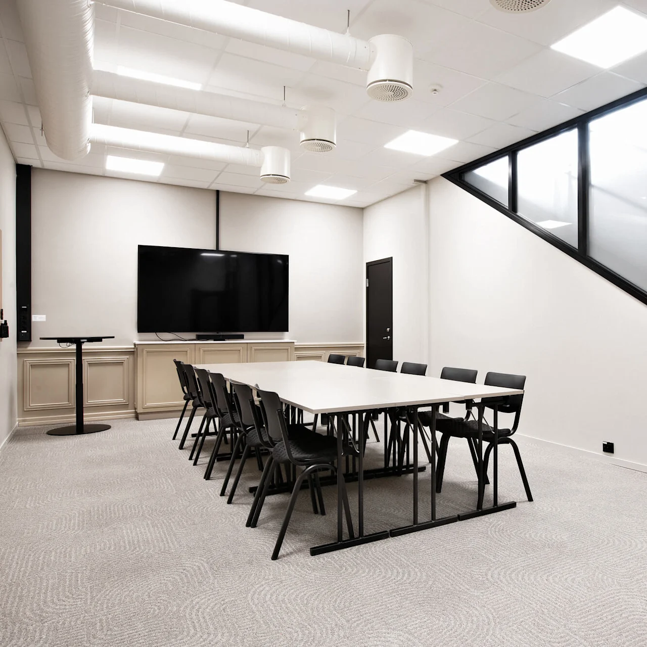 Et langt bord omgivet af sorte stole står i et lysfyldt konferencelokale med en stor TV, vinklet glasvæg og loftskanaler.