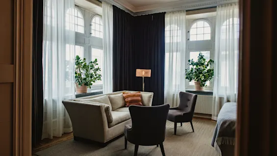 A beige sofa sits beside two gray chairs and a lamp on a carpeted floor. Large windows with sheer curtains and potted plants create a cozy, well-lit room.