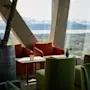 Hyggelige lænestole omgiver et lille bord ved store vinduer og tilbyder en naturskøn udsigt over en fjord og bjerge. Et potteplante står i nærheden og bidrager til den afslappede atmosfære.