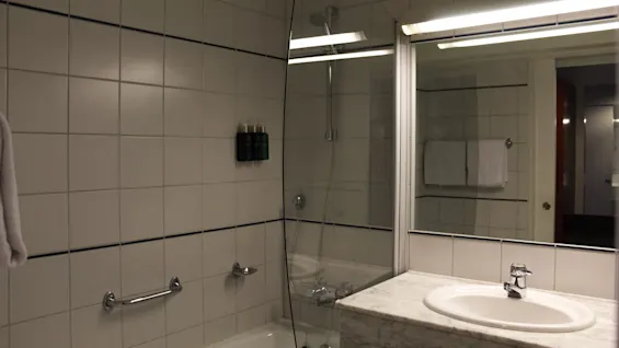 Et moderne badeværelse med flisebeklædte vægge, oplyst spejl over en marmorvask, og en bruseniche afgrænset af glas.