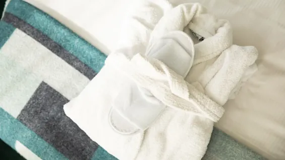 En mjuk vit morgonrock och matchande tofflor ligger prydligt på en bäddad säng, dekorerad med en geometriskt mönstrad blå och grå filt.