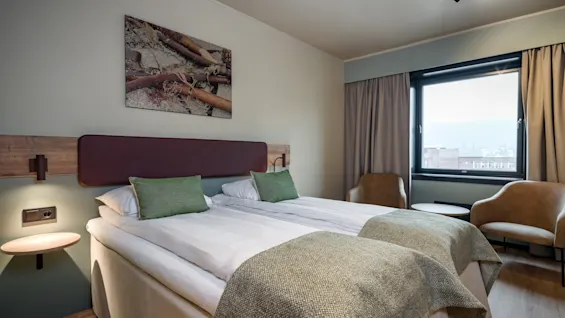 Et komfortabelt hotellrom med to senger, et stort vindu og to lenestoler, som antyder et avslappende opphold.