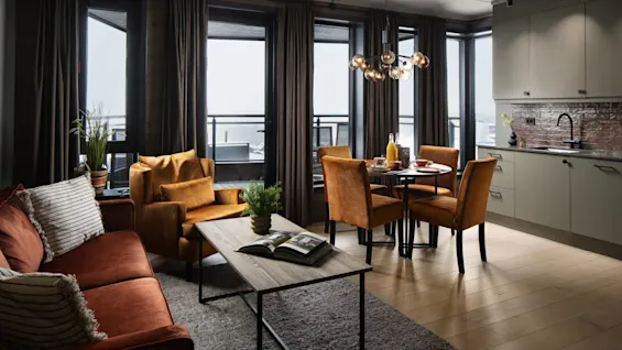 En hyggelig stue har en rustfarvet sofa og stole, et træbord med planter, ved siden af et spiseområde og moderne køkken, med store vinduer der vender ud mod en altan.