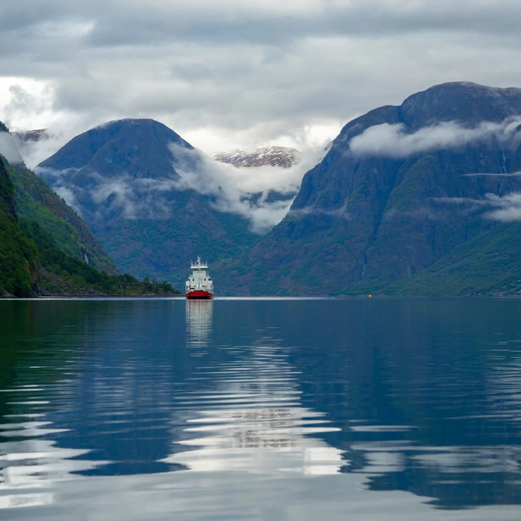 Vene purjehtii majesteettisen vuonon läpi Bergenissä, jota ympäröivät pilvien osittain peittämät vuoret.