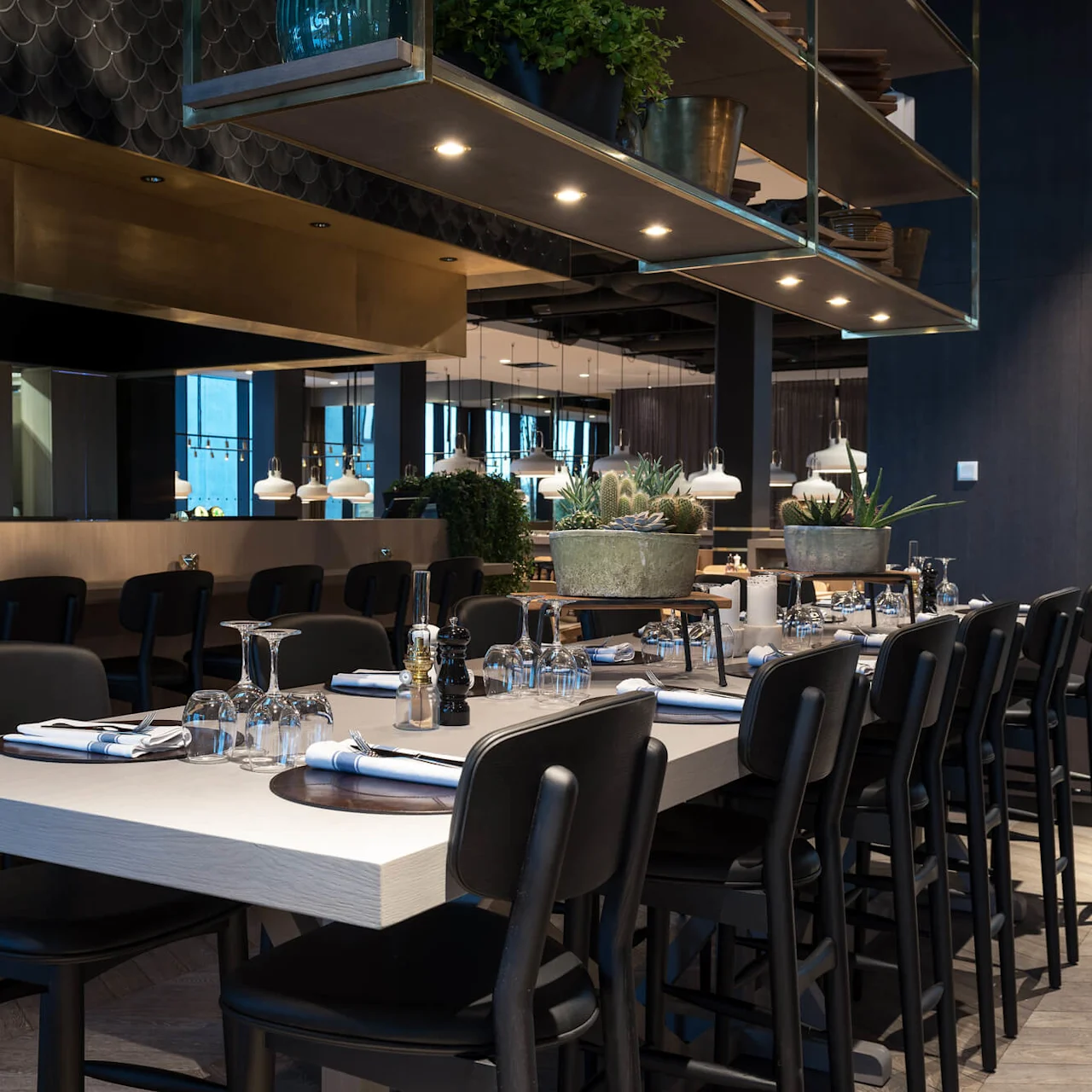 Bord dekket med glass og servietter, omgitt av svarte stoler. Over henger hyller med potteplanter. Moderne restaurantinteriør med dempet belysning og hengende lamper i bakgrunnen.