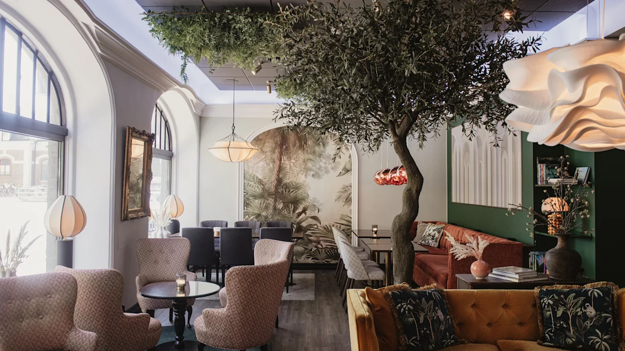 Ett träd dekorerar ett stiligt, mysigt café med bekväma sittplatser, eleganta lampor och stora välvda fönster som släpper in naturligt ljus. En muralmålning som föreställer naturen bidrar till den inbjudande atmosfären.