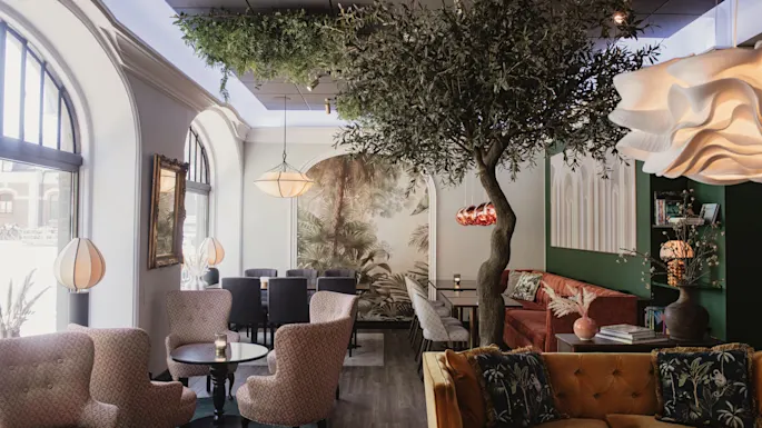 Et tre pynter en stilfull, koselig kafé med komfortable sitteplasser, elegante lamper og store buede vinduer som slipper inn naturlig lys. Et veggmaleri av naturen gir en innbydende stemning.