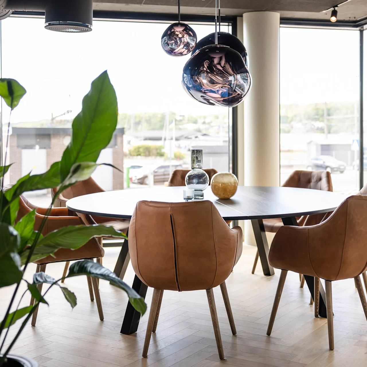 Et rundt bord med brune stole står i et lyst lokale med store vinduer, omgivet af dekorative hængelamper og indendørs planter.