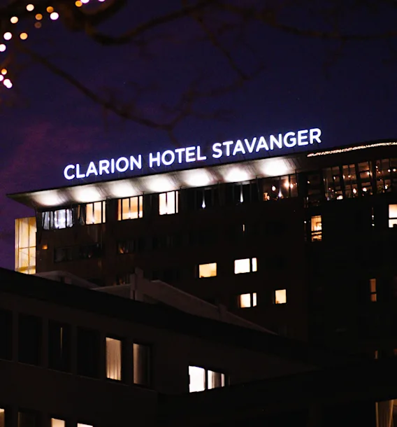 En hotellbyggnad på natten med 'HOTEL STAVANGER' upplyst på taket, inramad av ett träd med glittrande ljusslingor.