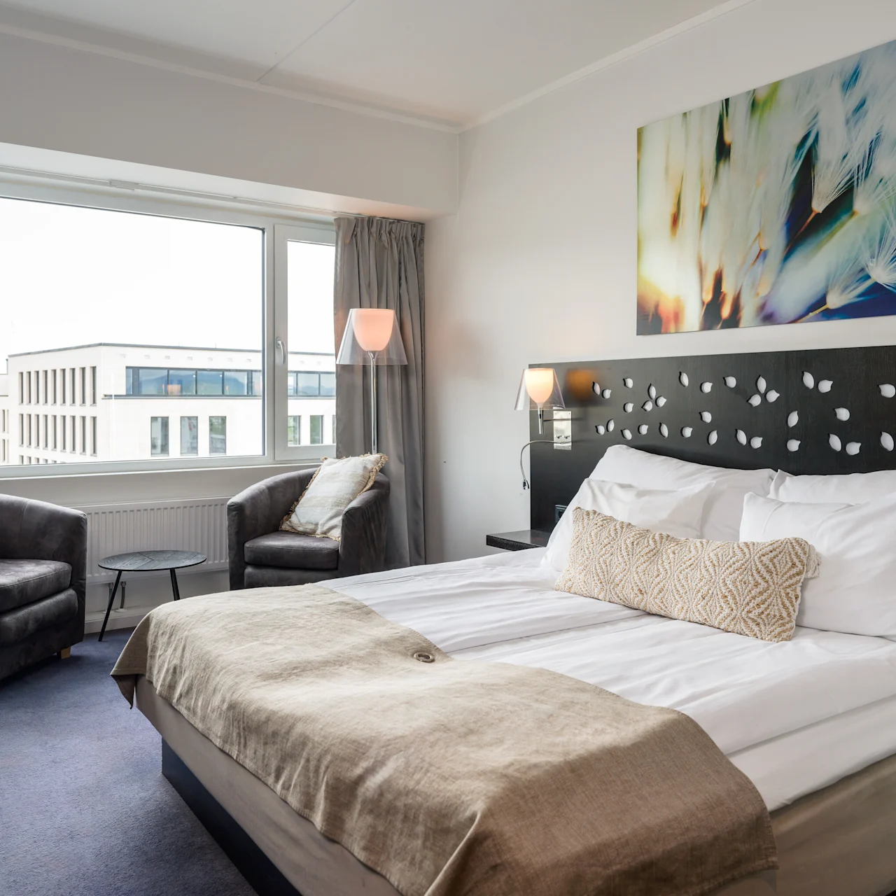 A neatly made bed with white linens and beige blanket; surrounded by armchairs and table near a large window, artwork above the headboard in a bright, modern hotel room.