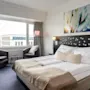 A neatly made bed with white linens and beige blanket; surrounded by armchairs and table near a large window, artwork above the headboard in a bright, modern hotel room.
