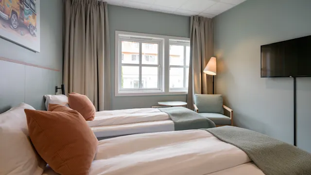 To pent oppredde enkeltsenger med brune puter, ved siden av store vinduer, et lite bord og en lenestol under en tent gulvlampe; en TV montert på en pastellblå vegg.