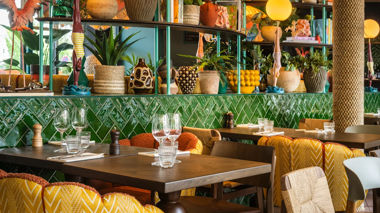 Borde dækket med glas og bestik, omgivet af farverige stole i en livlig restaurant. Dekorative planter og mønstrede fliser pryder baggrunden og skaber en livlig, eklektisk atmosfære.
