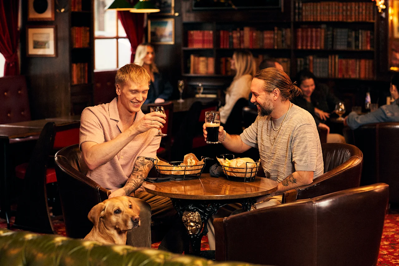 Kaksi ystävää nauttii juomia ja välipaloja viihtyisässä baarissa koiran kanssa. He nauravat ja pitävät hauskaa.