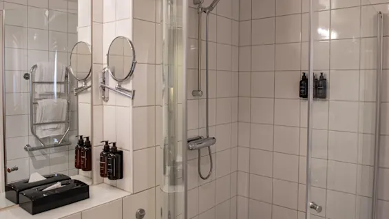 Shower with glass doors and wall-mounted toiletries in a tiled bathroom. Two round mirrors hang near a counter with a tissue box and more toiletries, adjacent to folded towels.
