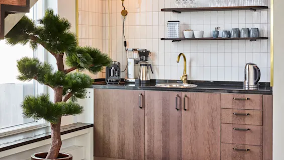 Et tremøbel støtter en svart benkeplate med vannkoker og kaffetrakter; hyller holder kopper og glass. Et potteplante står ved siden av, og en trapp er synlig.