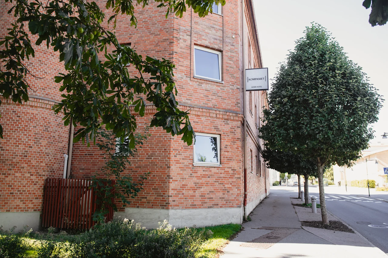 En tegelbyggnad med en skylt där det står KOMPANIET HOME HOTEL, omgiven av gröna träd och en trottoar.