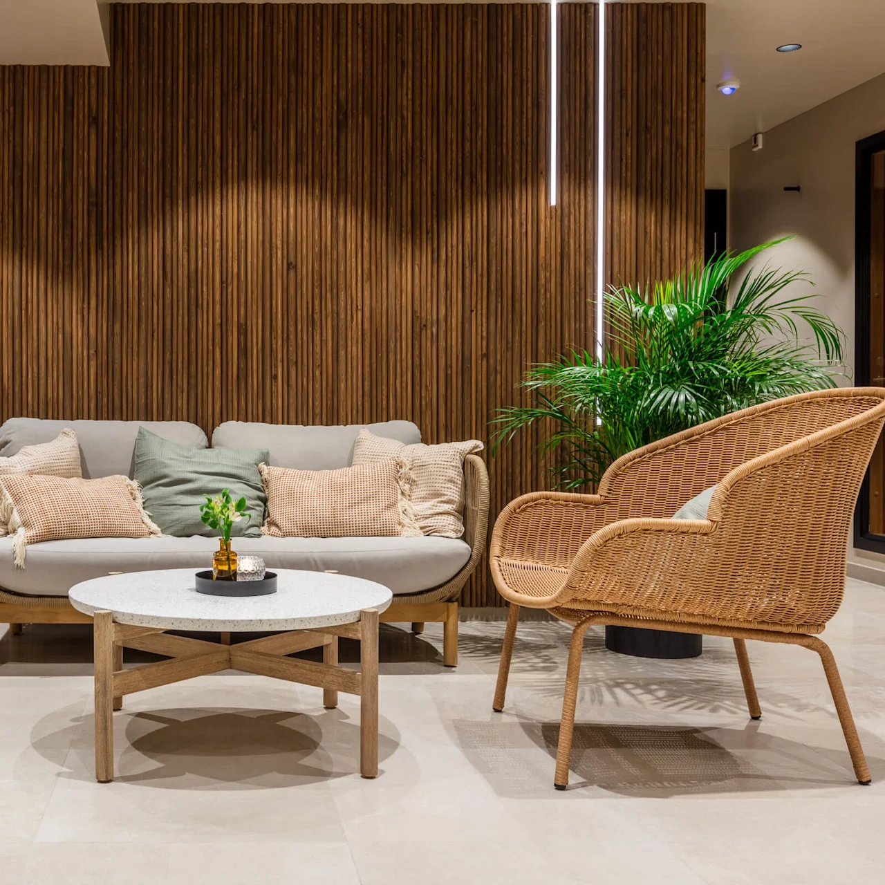 A modern living room with rattan armchairs, a sofa with cushions, and a coffee table, all on a tiled floor against a wooden slat wall with vertical lights.