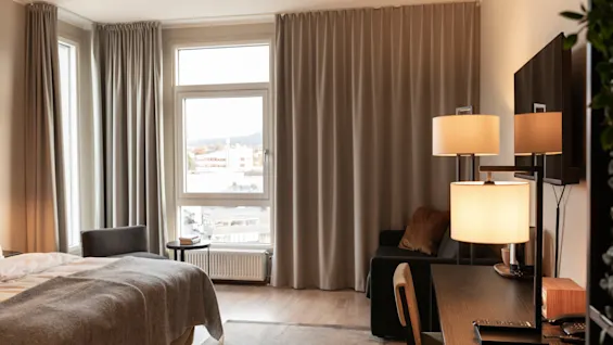 Et lyst og indbydende hotelværelse med en komfortabel seng, et funktionelt skrivebord og et stort vindue med udsigt over byen.