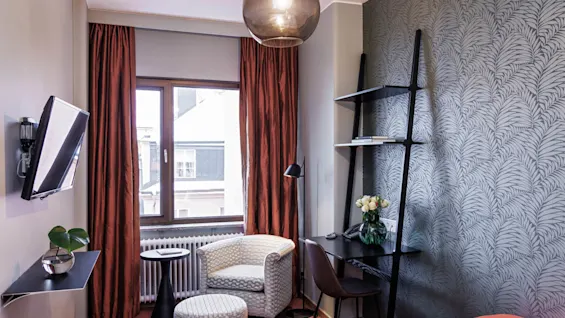 Ett stilfullt hotellrum med mönstrad tapet, moderna möbler och ett fönster som ger naturligt ljus.