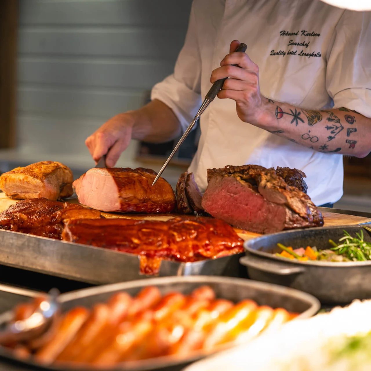 En kokk skjærer stekt kjøtt på et brett i et restaurantkjøkken. Tekst på kokkens jakke: "Richard Karlsson, Souschef, Quality Hotel Lappland."