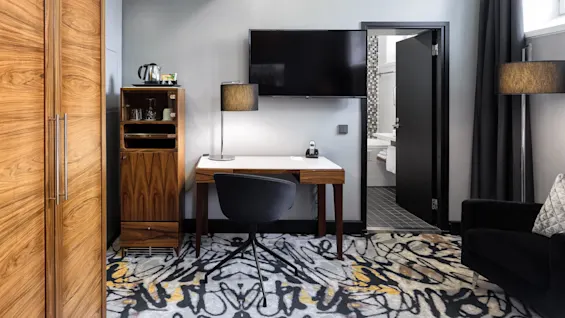 A modern hotel room with a desk, chair, and flat-screen TV; a minibar cabinet and open bathroom door reveal a neat, tiled interior.