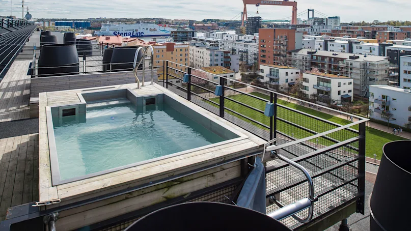 A rooftop pool with a metal ladder overlooks a cityscape, including residential buildings, a green park, and a large crane labeled "ERIKSBERG." A ferry with "Stena Line" visible in the background.