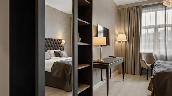 Et moderne hotelværelse med seng, skrivebord, TV og et hyggeligt siddeområde ved vinduet, adskilt af en reol.