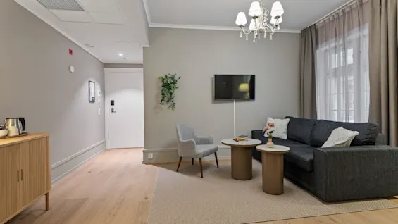 Et hyggeligt hotelopholdsområde med en mørk sofa, lys lænestol, sofaborde, et vægmonteret TV og en lysekrone.