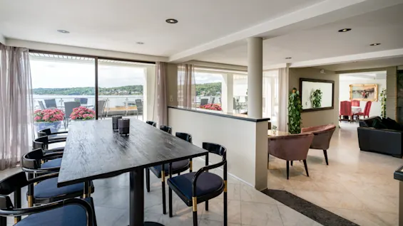 Et romslig spise- og salongområde med et langt bord og stoler, med utsikt over en innsjø og en balkong med blomster.