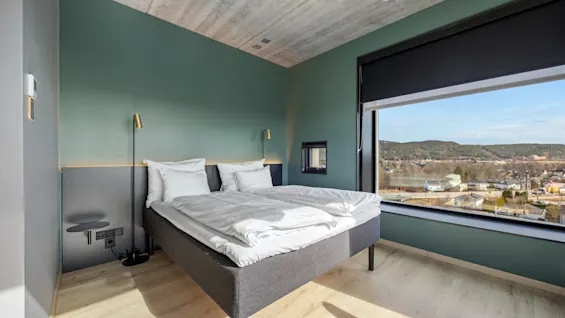 Et moderne hotellrom med en komfortabel seng, grønne vegger og et stort vindu med utsikt over en by.