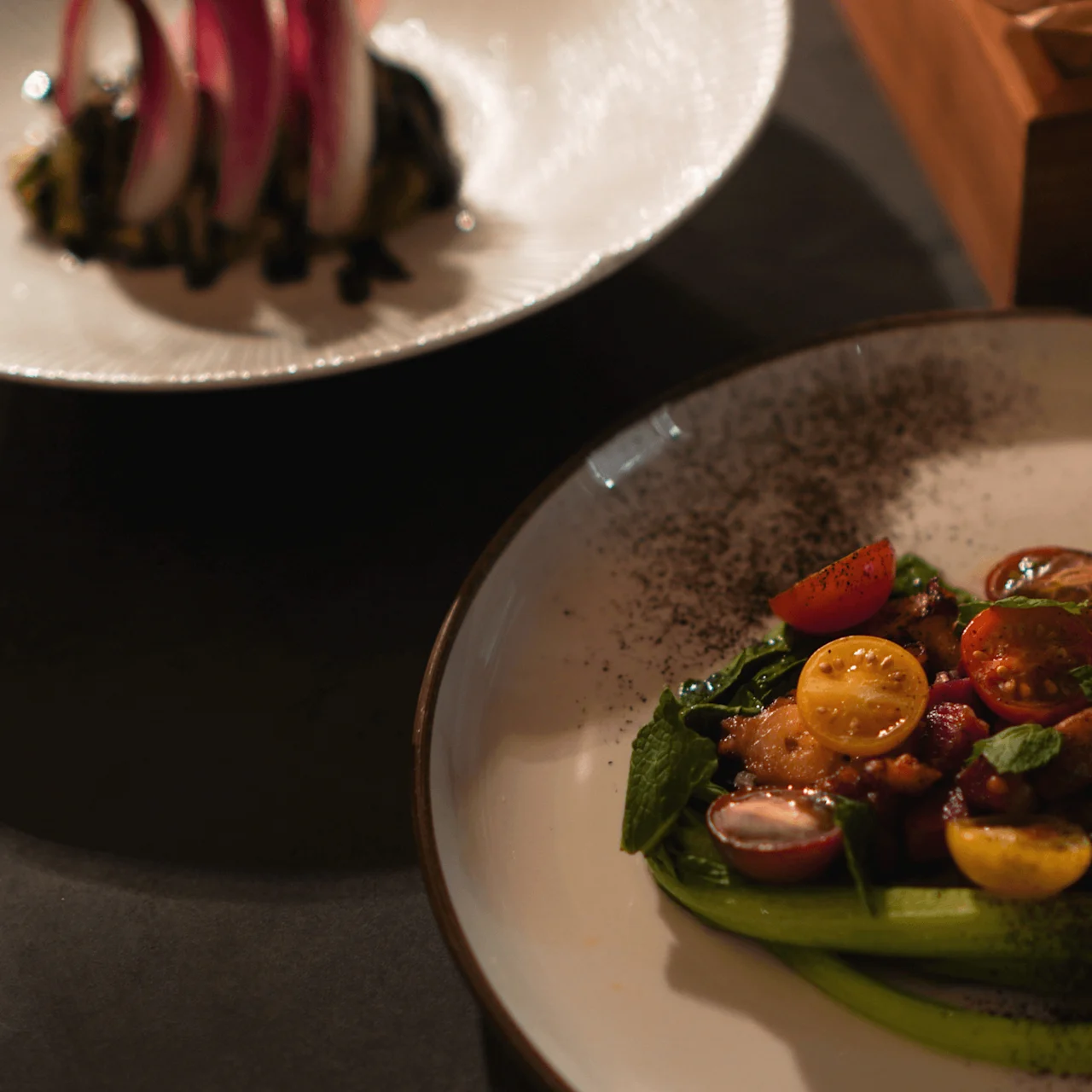 A plate holds a colorful dish with cherry tomatoes, greens, and dark seasoning, positioned beside another artistic food arrangement on a dark dining table. 