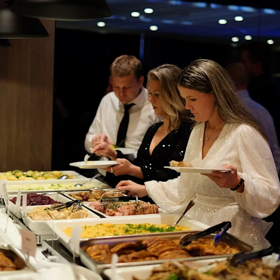 Folk forsyner seg med mat fra en stor buffet på et arrangement, med ulike retter tilgjengelig.